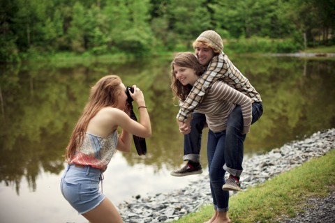 Girl photographing friend carrying boyfriend piggyback near lake