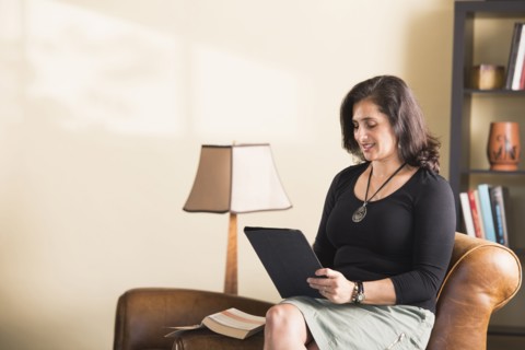 Hispanic woman sitting on armchair reading digital tablet