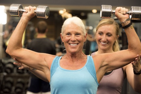 Trainer assisting older woman lifting dumbbells in gymnasium