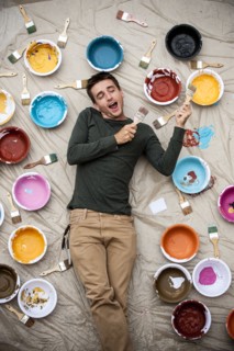 Man laying on tarp holding paintbrushes and singing
