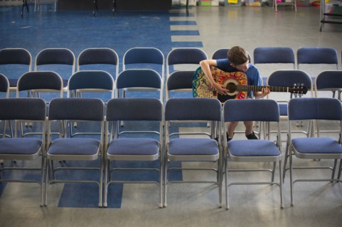 Caucasian boy sitting in row of chairs practicing guitar