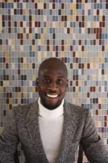 African man in tweed jacket smiling in front of tiled wall