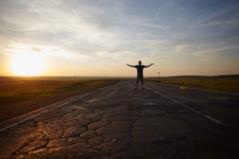 Caucasian man standing in cracked road at sunset with arms outstretched