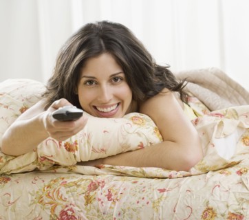 Hispanic woman laying on bed pointing remote control