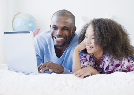African father and daughter using laptop on bed