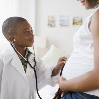 Black boy in lab coat holding stethoscope to pregnant mother's stomach