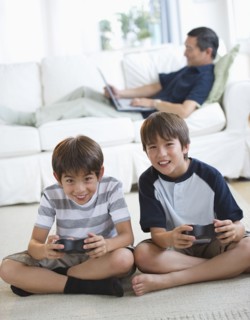 Smiling twin brothers playing video game while father uses laptop on living room sofa