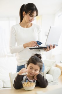 Hispanic mother shopping online while daughter eats cereal