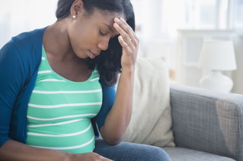 Anxious Black pregnant woman rubbing forehead on sofa