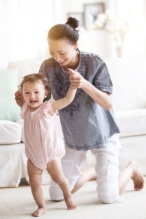 Mother helping baby daughter walk in living room