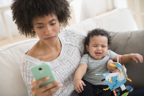 Mother holding crying baby son while texting on cell phone