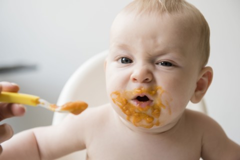 Mother feeding messy baby son with spoon in high chair