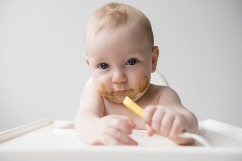 Messy Caucasian baby boy holding spoon in high chair