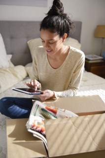 Hispanic woman sitting on bed near box using digital tablet