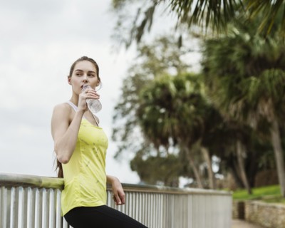 Woman in sports clothing leaning on fence and drinking water from bottle
