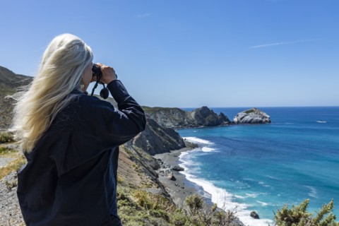 Rear view of senior blonde woman looking at ocean with binoculars on Big Sur coast