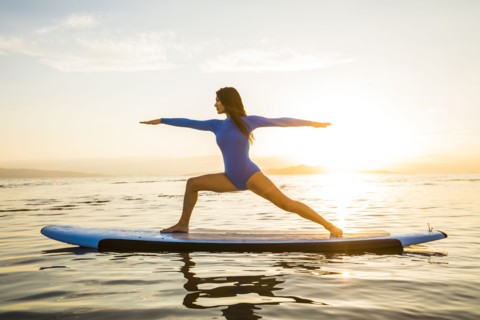 Woman in blue swimsuit performing Virabhadrasana II (Warrior II Pose) on paddleboard at sunset