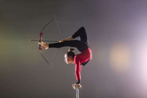 Young acrobat performing with archery bow on gymnastics bar