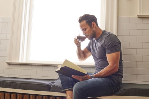 Man reading book and drinking tea by window at home