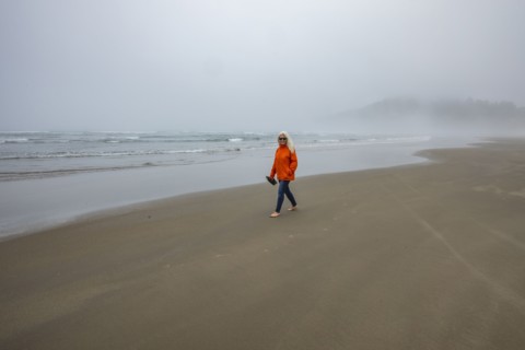 Woman in orange jacket walks along foggy beach