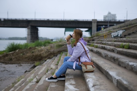 Thoughtful woman sitting on steps and drinking coffee while looking at river