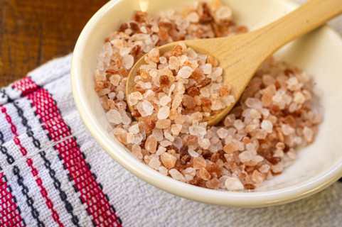 Coarse pink salt crystals in white bowl on kitchen table cloth and wooden spoon