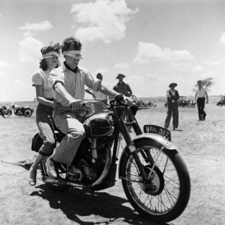 Blindfolded biking. A blindfolded couple sit astride a motorcycle at a motor sports gymkhana. Pat