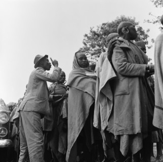 Queuing at a cleansing ceremony. A man with a raised finger speaks over the top of a crowd of women
