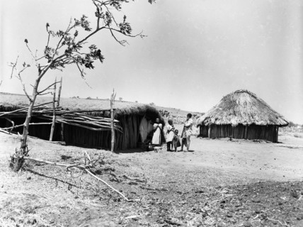 Mbulu hut. An adult and three children stand outside a long wooden hut with a flat, thatched roof.