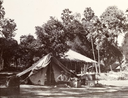 Camping in a village. A European man sits on an upturned crate outside his tent, brandishing a