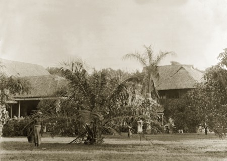 In the garden of a colonial bungalow. A barechested servant carries a bucket across the lawn of a