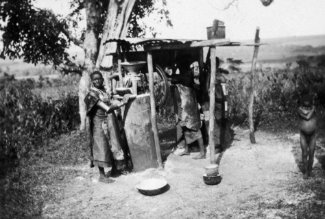 Nandi women grinding maize. Two Nandi women operate a small, partially-mechanized maize mill, which