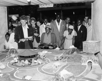 Driving game. A mixed race crowd gathers around a detailed miniature road model at the Motor Sports