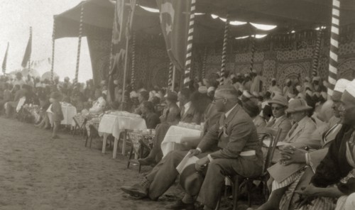 Kings' Day celebrations. A large, mostly male, crowd of onlookers sit around tables beneath a