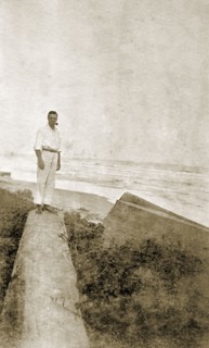 Mr Tamlin on a log. Mr Alfred Tamlin stands on a fallen log on a cliff overlooking the coast.