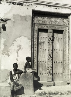 Carved wooden door, Zanzibar. A young man and a child sit in the sunshine beside an ornately carved