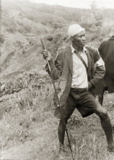 A Kikuyu Home Guard on duty. A young man in a tattered jacket stands barefoot on a hillside,