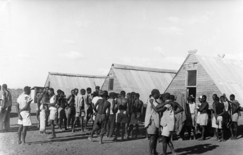 Home Guards at their living quarters. Young Kikuyu men gather beside prefabricated huts, a
