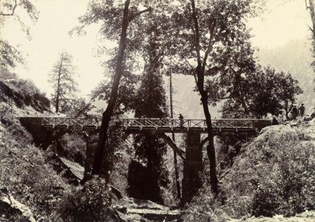 Bridge on the way to Kashmir. A distant figure peers over the side of a wooden road bridge,