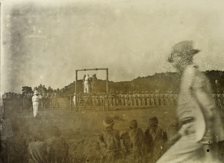 Colonial execution. An executioner prepares for two African men to be hung from a wooden scaffold,