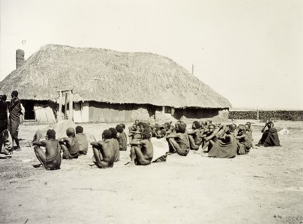 Depot for hiring porters. A group of African men and boys sit outside a large, thatched building,