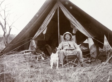 Frederick Stanbury outside a tent. Stanbury is seated on a canvas chair in front of a small open