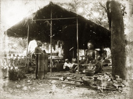 Idol-making, Burma (Myanmar). Two craftsmen sit, under a thatched, open-walled shelter, burnishing