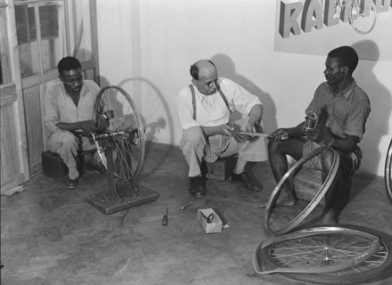 Kassam Kanji's workshop'. Two Kenyan men work alongside a European, repairing bicycle wheels in a