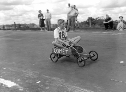 Number 18 in the soap box derby. An uneasy-looking boy rides to the finish line in his homemade
