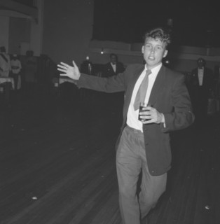 On the dance floor. A young man dressed in a suit approaches the camera, glass in hand, from across