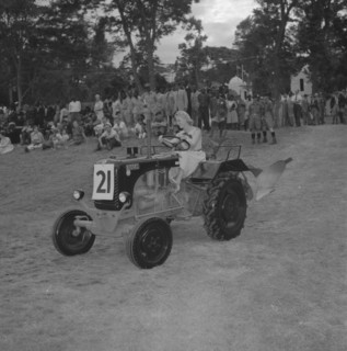 Tractor in a vehicle parade. A young woman with coiffeured hair and heels, drives a small tractor