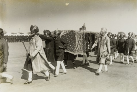 Maharajah in a sedan chair. A procession of attendants dressed in ceremonial attire, carry a sedan