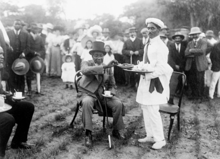 Tea and cake at a royal reception. Dressed in a Western-style suit and top hat, an African chief