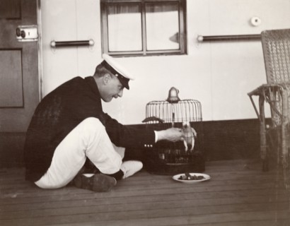 Caged parrot aboard the S.S. Balmoral Castle. A British naval officer sits cross-legged on the deck
